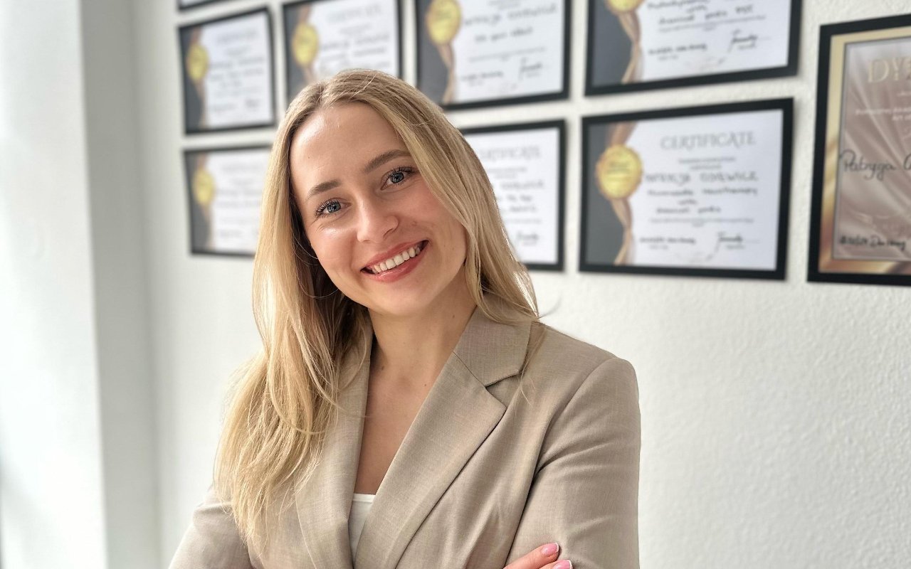 Patrycja, glimlachend in haar salon, met ingelijste certificaten aan de muur achter haar